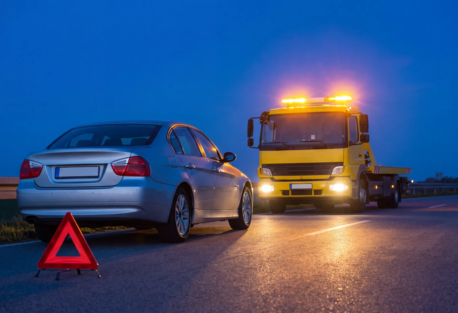 Pannenhilfe im Nachteinsatz Liegengebliebenes Auto bei Nacht mit Warndreieck und herannahendem gelbem Abschleppwagen mit leuchtender Rundumkennleuchte